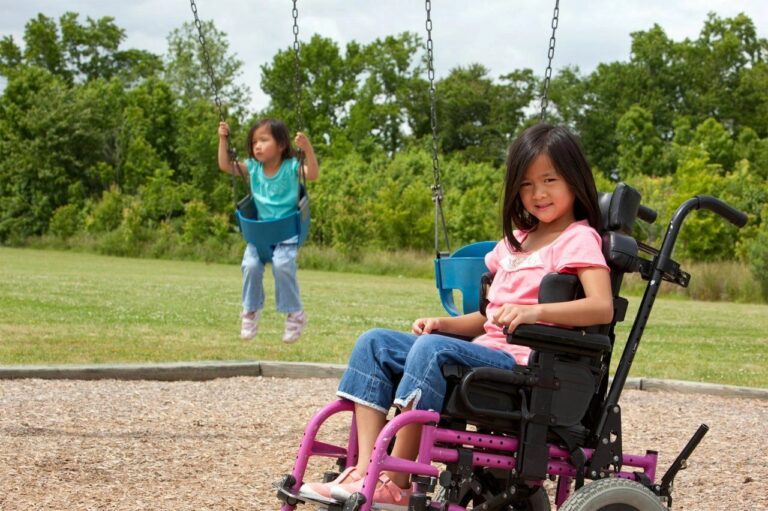 A girl in a wheelchair watches another girl on the swings